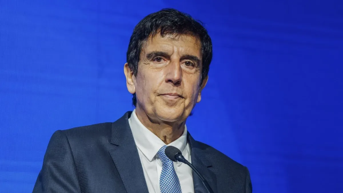 Carlos Melconian en una conferencia, con gesto serio y micrófono, fondo con banderas argentinas